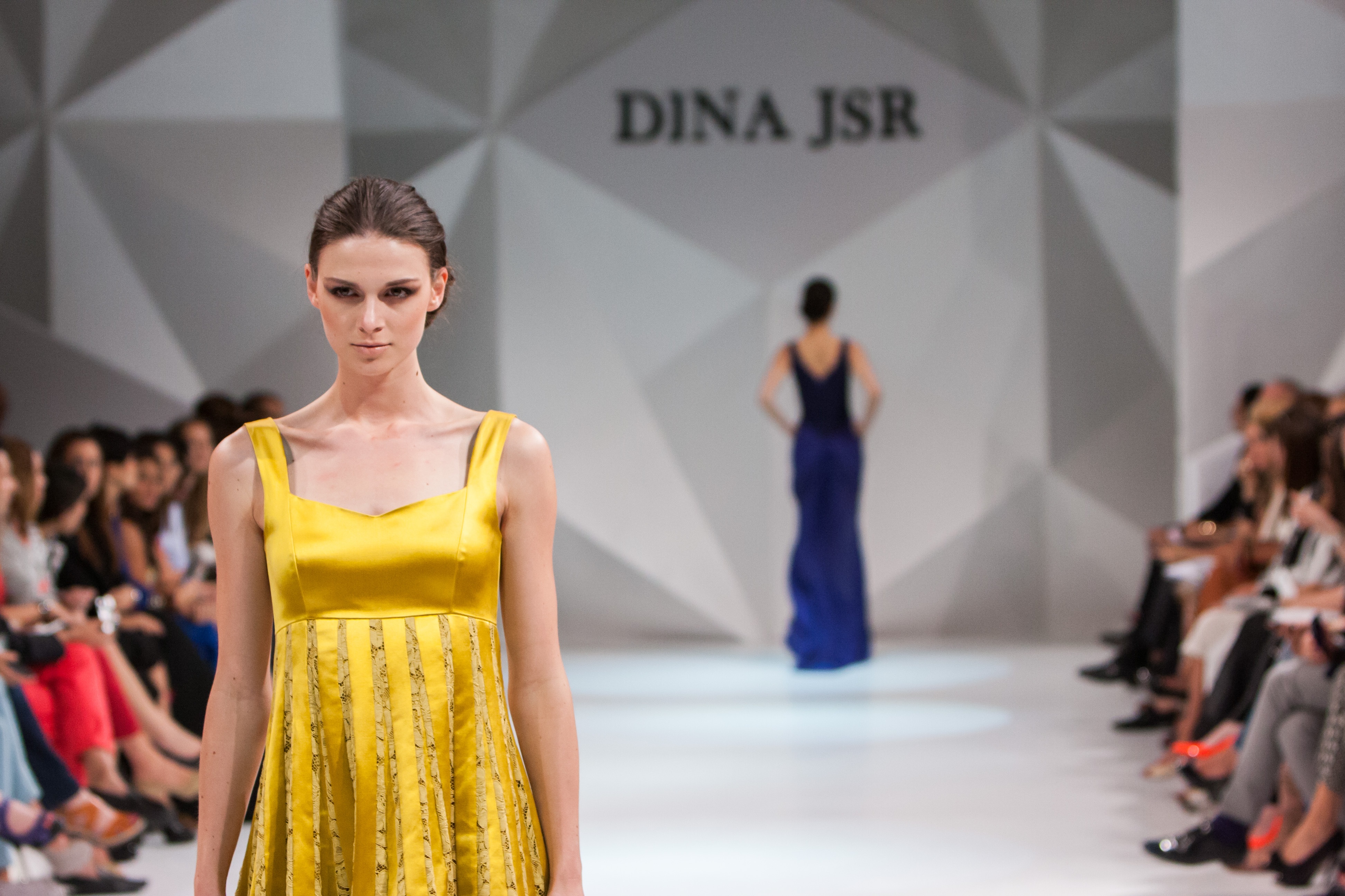 A fashion model walking a lit runway while spectators watch from both sides.